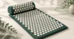 Mørkegrønn spikematte og tilhørende nakkepute med hvite plastpigger på et lyst underlag.