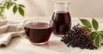 En glasskopp med dampende svarthyllsaft og en karaffel står ved siden av modne svarthyllbær og grønne blader på et lyst bord.